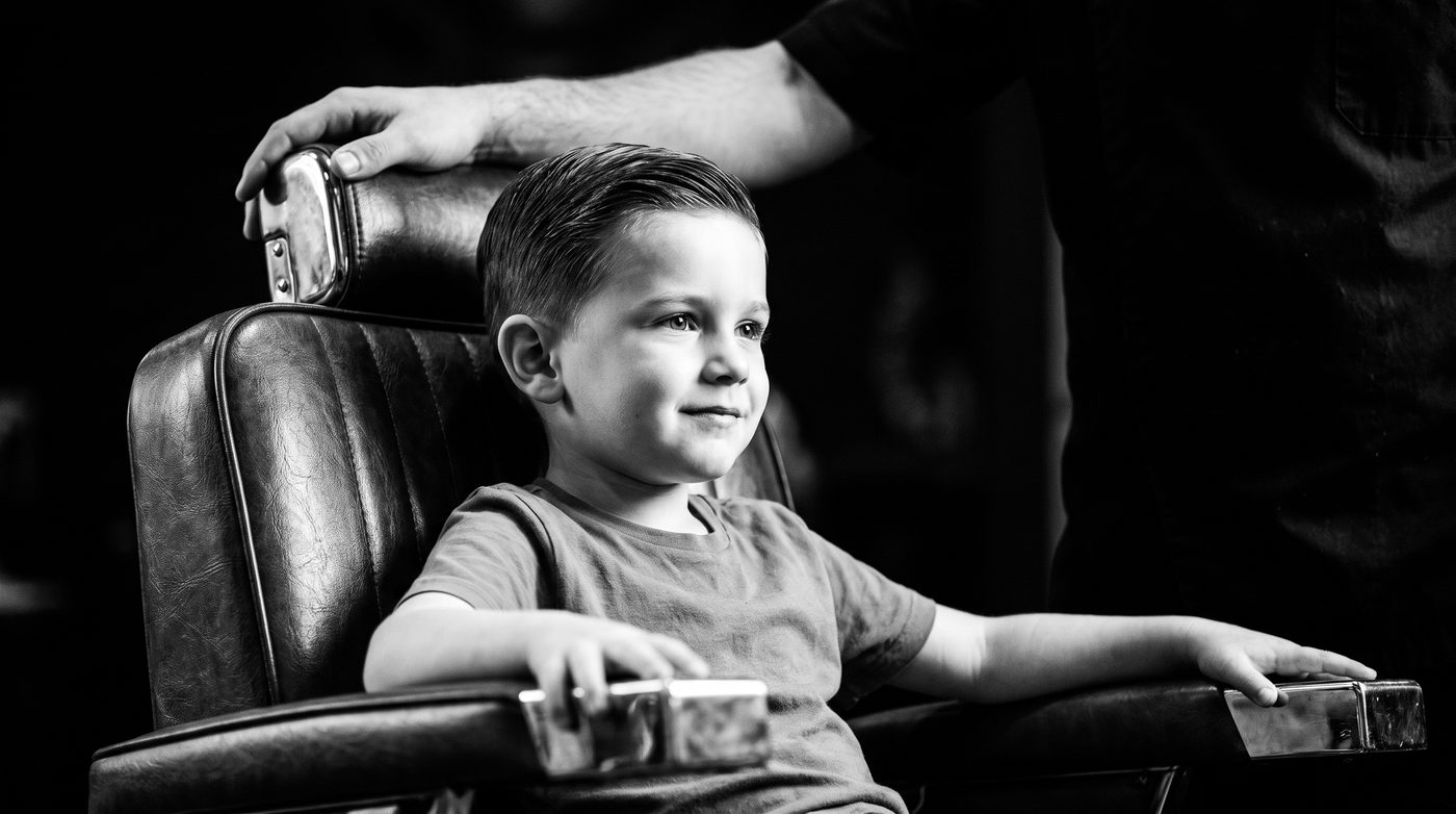 A young boy sitting up straight and proud in a vintage leather barber chair, a barber’s hand resting on the chair back